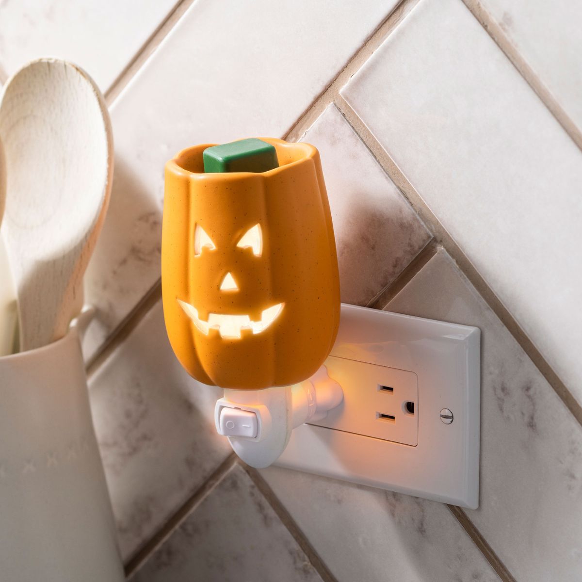 Pumpkin-shaped fall wax warmer plugged into a wall socket on a tiled wall.