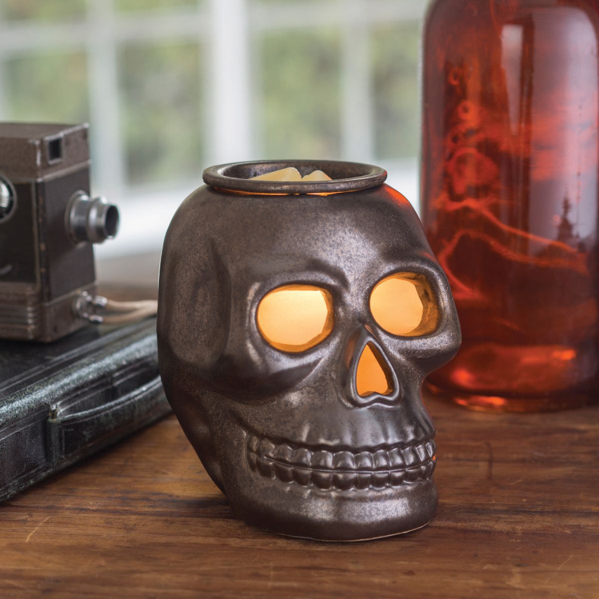 Skull-shaped wax warmer with glowing interior on a wooden surface, with a camera and glass bottle in the background