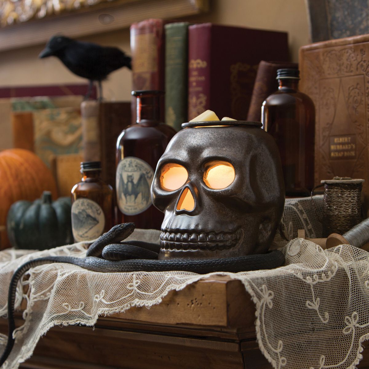 Skull-shaped wax warmer with glowing eyes on a table with pumpkins, books, and bottles in the background.