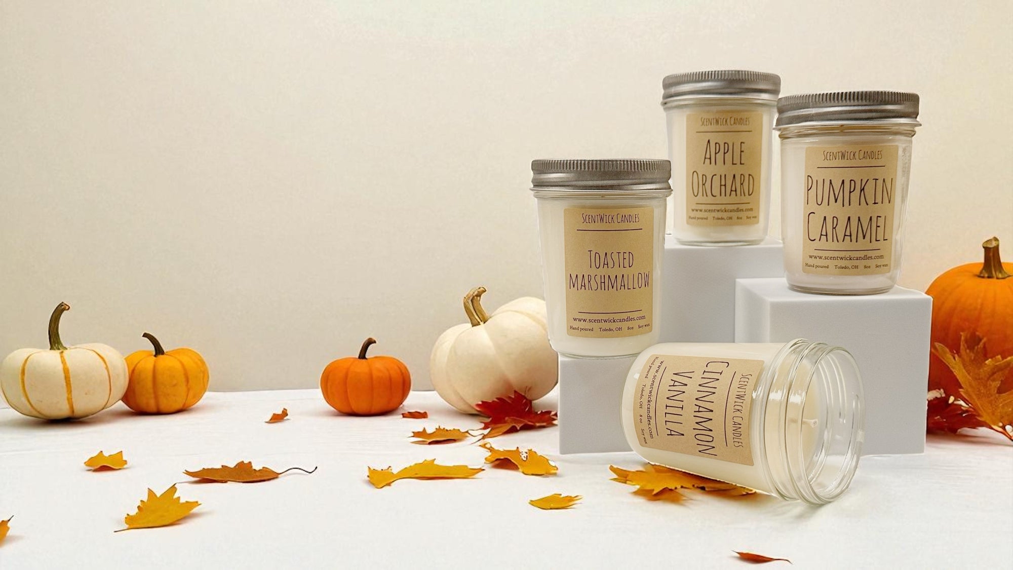 Collection of fall candles with labels on a white surface with pumpkins and leaves.