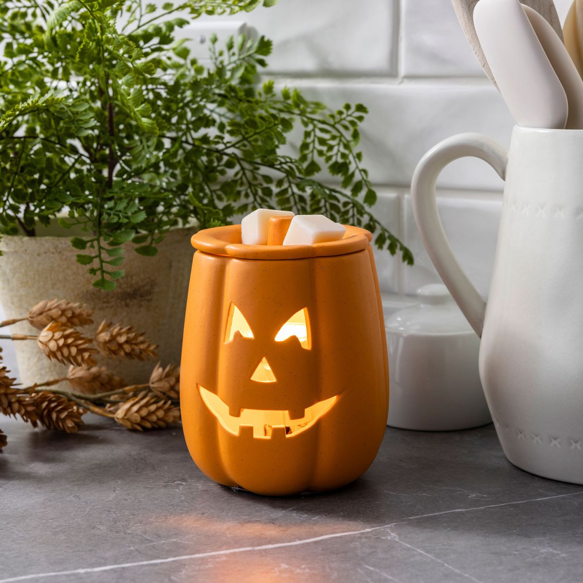 Orange jack-o'-lantern-shaped wax melt warmer on a surface with decorative elements.