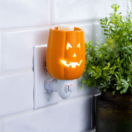 Orange jack-o'-lantern wax warmer plugged into a wall socket with a plant in the background.