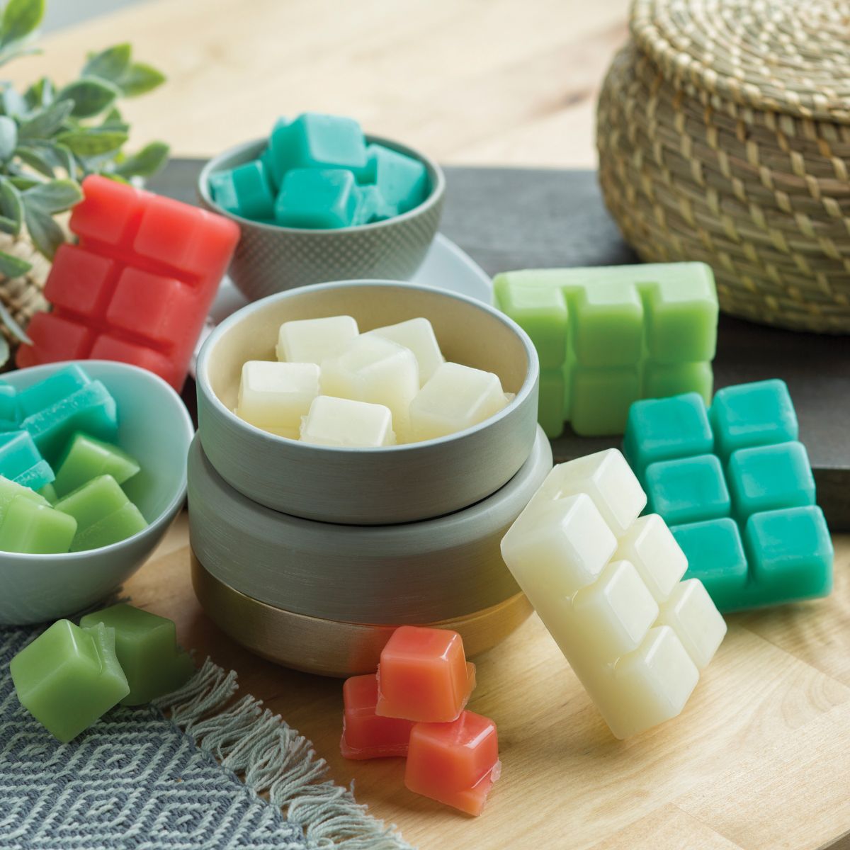 Colorful wax melts in small bowls on a wooden surface with a plant and woven basket in the background.