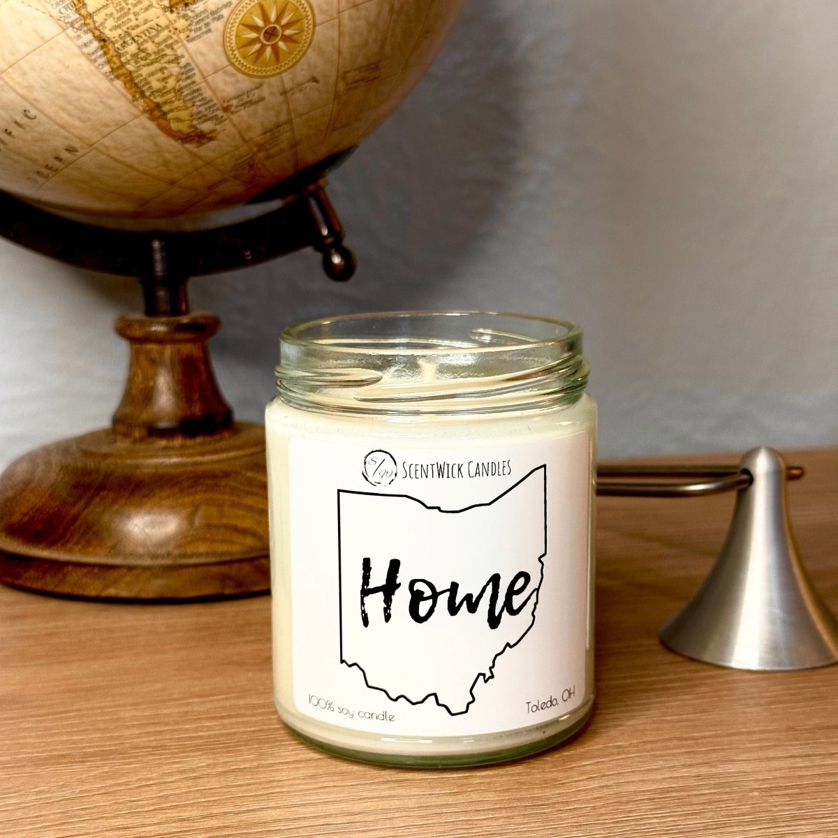Candle jar labeled 'Home' with an Ohio map design next to a wooden globe on a wooden surface.