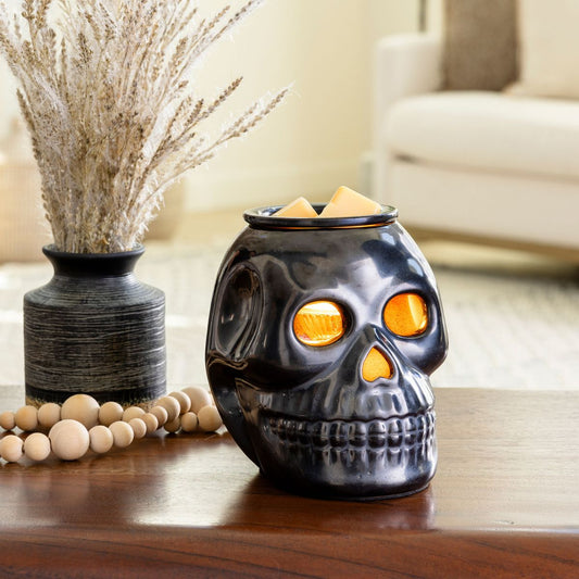 Skull-shaped wax warmer with glowing eyes on a wooden surface, surrounded by decorative items.