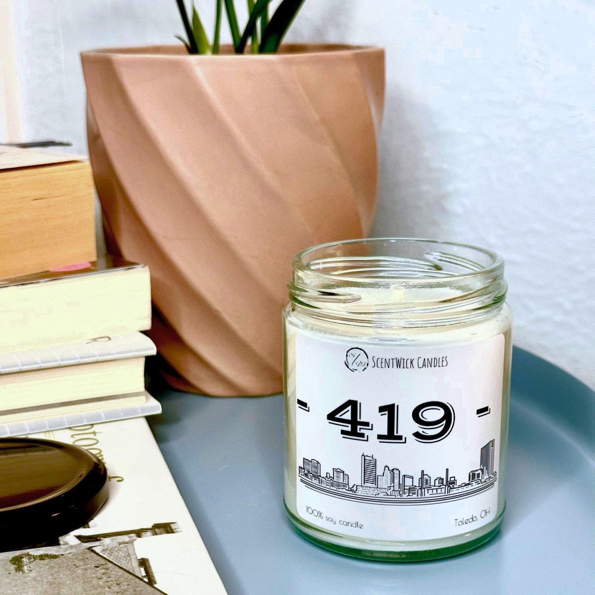 Candle in a glass jar with a label featuring the number '419' on a surface with books and a plant in the background.