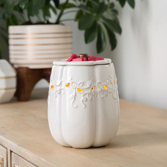 White ceramic pumpkin wax warmer with floral design on a wooden surface, surrounded by potted plants.