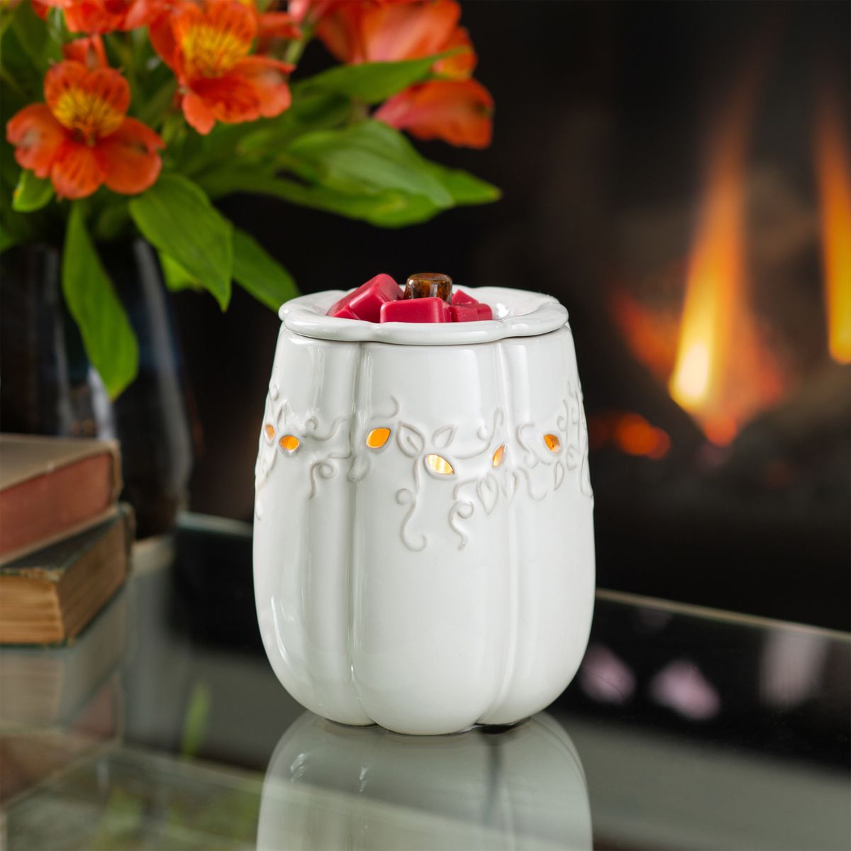 White ceramic pumpkin wax warmer with decorative elements on a reflective surface in front of a fireplace.
